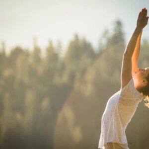 Frau bei einer Yoga-Dehnung mit nach oben gestreckten Armen im Sonnenlicht vor einem Wald.