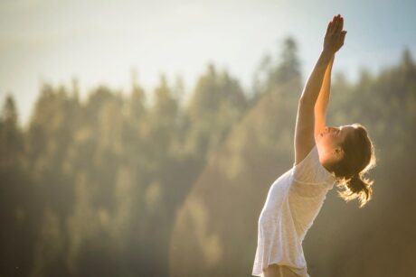 Frau bei einer Yoga-Dehnung mit nach oben gestreckten Armen im Sonnenlicht vor einem Wald.